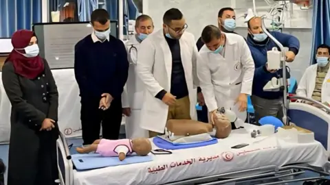 Handout Masked medical students gather around a plastic dummy on a gurney to watch a demonstration. A baby doll is also on the bed.