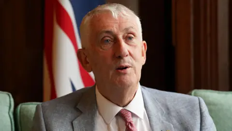 PA Media Sir Lindsay Hoyle is speaking to the camera while seated in a green sofa and wearing a grey suit jacket and a salmon pink tie.