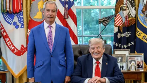 @Nigel_Farage Nigel Farage stands beside a seated Donald Trump in the Oval Office. Farage wears a blue suit with a purple tie, while Trump is in a dark suit with a red tie. They are behind the Resolute Desk, which holds phones, documents, and other items. The room features large windows showing greenery outside, with American and military flags, statues, and framed photos decorating the space.