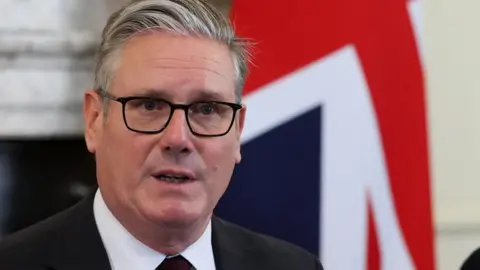 PA Media Keir Starmer is pictured in front of union jack, wearing a black suit with a dark red tie and glasses with his mouth slightly open, his face stern.