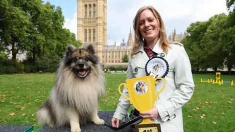 Kieran Cleeves/PA Media A small fluffy dog with dark brown fur around its face and cream fur on its body smiles at the camera. He is sitting on a grey mat with his owner, a blonde-haired woman in a white coat, standing next to him. She has a white rosette pinned to her coat and is holding a yellow-coloured cardboard cut-out of a trophy. They are on a grassy area in front of the Houses of Parliament.